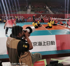 João Maia fotografando jogo de volei feminino paralimpico em Tóquio. Na cena há cinco atletas do Brasil disputando a partida com o time do Canadá.