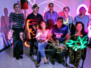 João maia no centro da imagem, trajando camisa azul com logo da fotografia cega, calça jeans azul, 9 pessoas com e sem deficiência, reunidas ao redor da imagem.