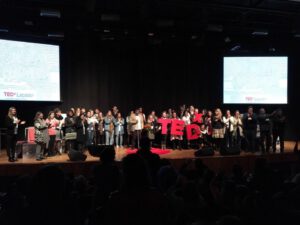 João maia no centro da imagem, trajando camisa social branca, blazer na cor caramelo, calça jeans preta, pessoas reunidas ao redor da imagem, segundo plano telão com a logo TEDx lançador.