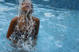Retrato por João Maia de mulher que está emergindo do meio da piscina. Negra usando maiô. A agua que está caindo no seu corpo forma uma imagem abstrata. A piscina tem fundo azul