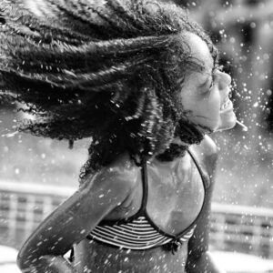 foto em preto e branco na horizontal, menina, cor da pele parda, cabelos pretos crespos, sorriso largo, ela brincar numa piscina Geraldo sua cabeça junto com seus cabelos seus cabelos crespos ao vento, e gotículas de água.