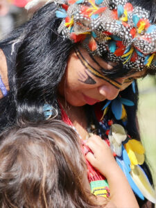 Retrato de uma índia da tribo Guarani abraçando sua filha. A índia está de perfil com a cabeça abaixada. Usa penas coloridas na cabeça e também penas pretas com bolinhas brancas. Possui linhas pretas que contornam o rosto. Também usa brinco de penas azuis e amarelas. Usa um colar de miçangas vermelhas. A criança está de costas para a foto, porém é possível ver seu cabelo comprido e sua mãozinha que segura a gola da roupa da mãe