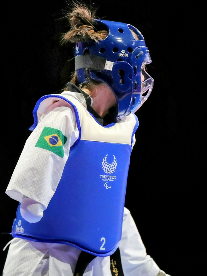 Atleta feminino de Taekwondo da seleção brasileira nas paralimpíadas de Tókio . Ela está de pé de perfil com kimono branco, protetor de cabeça azul, colete branco e azul com a palavra Tokio 2020 e bandeira do Brasil abaixo do ombro direito . Ela não possuí o antebraço e mão direita . Ela está com faixa preta . O segundo plano é todo preto