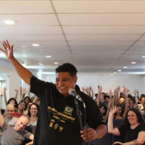 A imagem mostra João Maia, um homem sorridente em pé, falando ao microfone, com o braço direito levantado em um gesto de saudação. Ele está usando uma camiseta preta com a inscrição "Blind Photography" e outros textos menores. Atrás dele, há uma plateia de pessoas sentadas, muitas delas também com os braços levantados, aparentemente participando de uma interação ou saudação. O ambiente parece ser uma sala de conferências ou auditório, com teto branco e iluminação embutida. Todos parecem estar felizes e engajados no evento