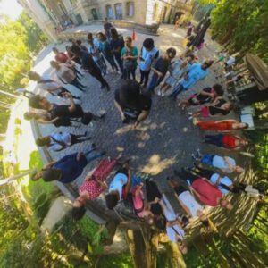 A imagem mostra um grupo de pessoas em um espaço ao ar livre, provavelmente em um parque ou jardim. Elas estão dispostas em um círculo, olhando para o centro, onde parece haver uma pessoa tirando uma foto ou filmando. O chão é de pedras e ao fundo há um edifício de pedra com janelas. Ao redor, há muitas árvores e vegetação, sugerindo um ambiente natural. A foto foi tirada com uma lente grande angular ou olho de peixe, o que dá um efeito curvado à imagem.