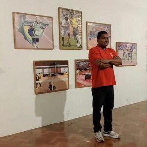 A imagem mostra João Maia,homem de pele morena, vestindo uma camisa polo vermelha e calça preta, com os braços cruzados, em pé em frente a uma parede branca. Na parede, há cinco quadros com fotos de esportes. - No quadro superior esquerdo, há um atleta segurando uma bandeira do Brasil. - No quadro superior central, dois jogadores de futebol, um deles vestindo a camisa da seleção brasileira. - No quadro superior direito, uma cena de um evento esportivo com várias pessoas. - No quadro inferior esquerdo, uma partida de vôlei sentado. - No quadro inferior direito, uma cena de um evento esportivo em um ginásio. O chão é de madeira e o ambiente parece ser uma galeria ou exposição de arte relacionada a esportes.