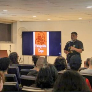 A imagem mostra uma apresentação em um auditório. No centro, oão Maia de pé, falando ao microfone. Ele está usando uma camiseta preta. À sua esquerda, há uma tela grande exibindo um slide com um fundo laranja e a imagem de um homem segurando uma câmera próxima ao olho. O texto no slide diz "Fotografia Cega". Na frente de João, há várias pessoas sentadas, assistindo à apresentação. O ambiente parece ser uma sala de conferências com iluminação de teto.