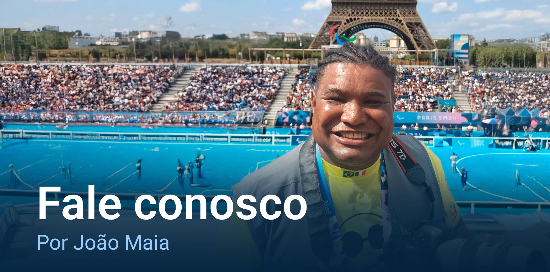 João Maia sorridente em frente à Torre Eiffel. Ele está vestindo uma camiseta amarela com um desenho de emoji símbolo da Fundação Dorina Nowil com rosto sorridente e óculos escuros, e um colete cinza. No pescoço, há um crachá com fitas coloridas e bandeiras do Brasil e de outro país. Ele segura uma câmera fotográfica grande. Ao fundo, há um campo de esportes azul com pessoas jogando e uma arquibancada cheia de espectadores. Acima da Torre Eiffel, há os anéis olímpicos e o símbolo paralímpico. O céu está azul com algumas nuvens. Há também banners com a inscrição "Paris 2024".