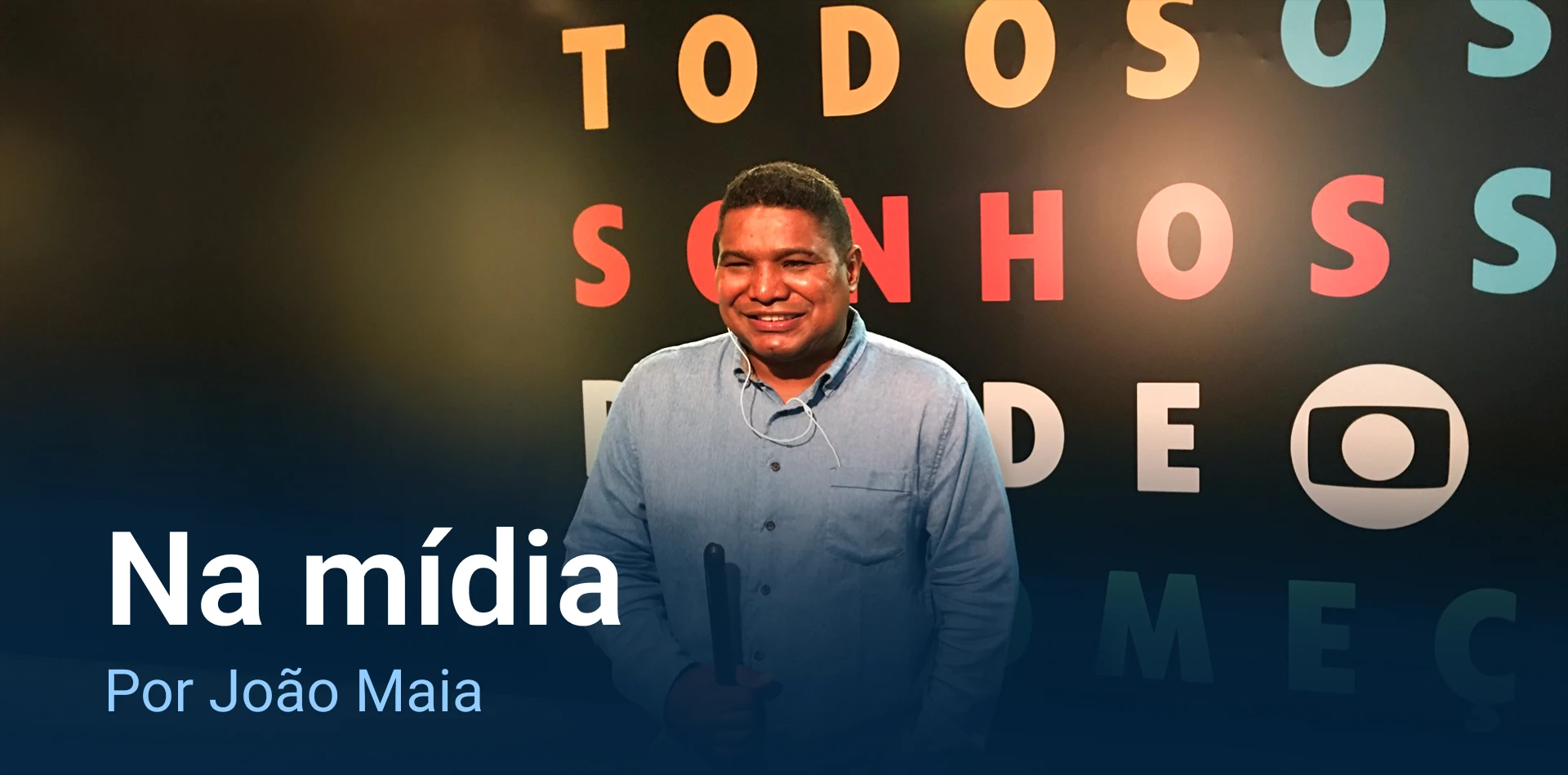 João Maia , negro , cabelo crespo preto ondulado , está com um sorriso contagiante. João veste uma camisa azul de manga longa, de pé, está com a mão esquerda no bolso. Com a direita segura a bengala. No plano de fundo símbolo da Globo num painel preto com letras coloridas formando a palavra " Todos os sonhos'
